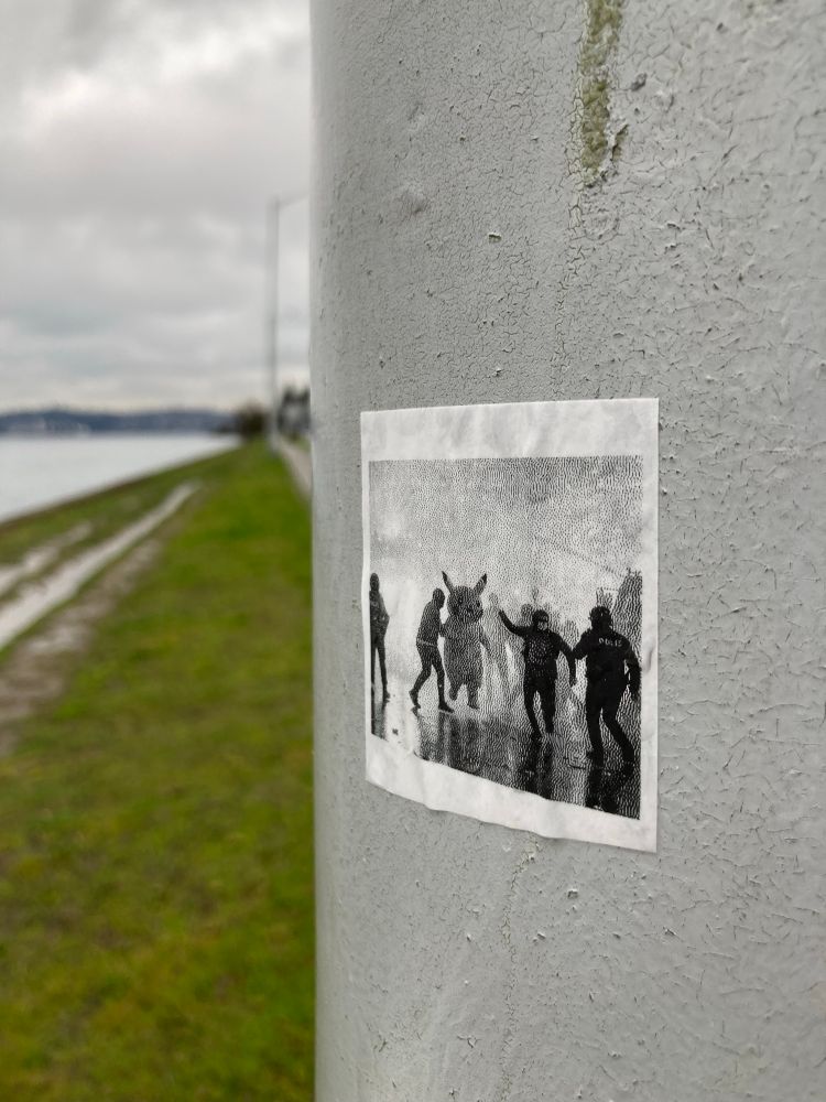 a square handmade label (?) sticker on a utility pole. it shows a black and white picture of a person in a pikachu blow up costume fighting the police.