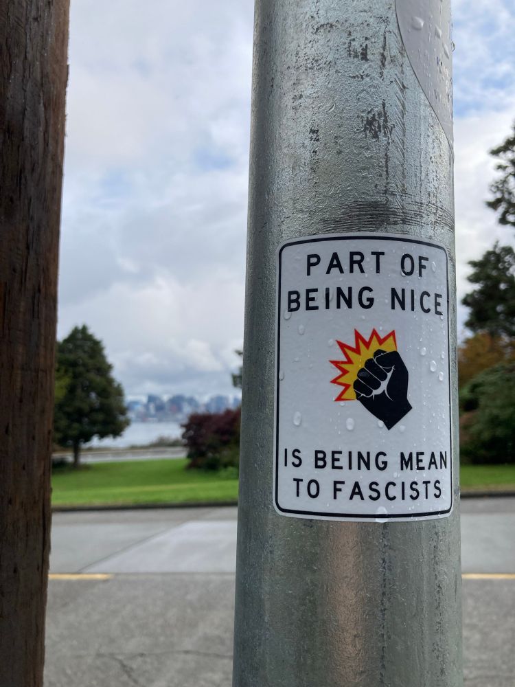 a sticker on a utility pole with a few raindrops on it. A city skyline out of focus and a park in the background. 

the sticker has a white background with a black outline that’s reminiscent of a street sign. in highway gothic font it say:
PART OF BEING NICE
IS BEING MEAN TO FASCISTS

in the center is a black power fist icon moving up and to the left. a “POW” cloud sits around it with yellow in the middle and a red outline to show some action. 