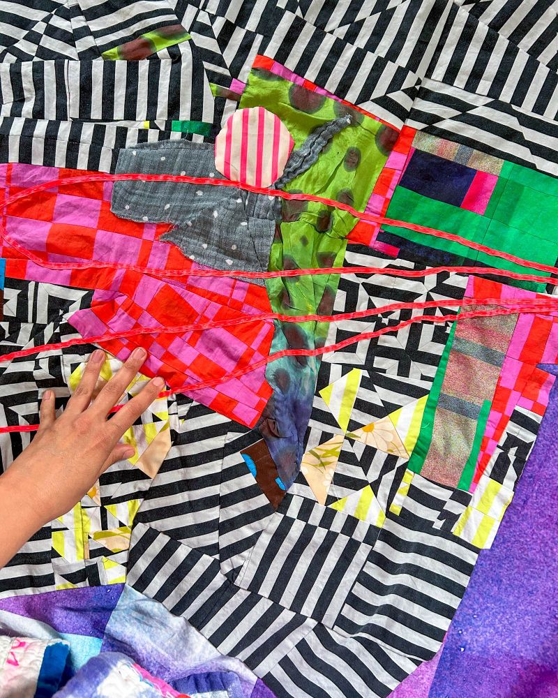 Detail of an abstract quilt top with black and white stripes on a purple-blue soft grid background with hand in bottom left hand corner. 