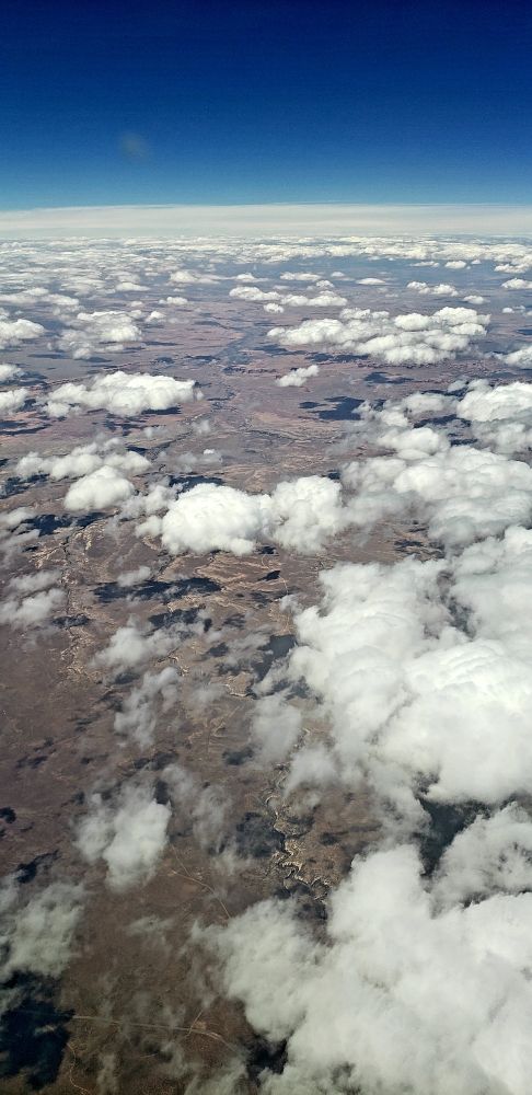 A picture of a taupe-colored desert from high altitude. White clouds dot the sky over the desert as they extend into the far distance. They end where the curved edge of the earth meets the bright blue sky