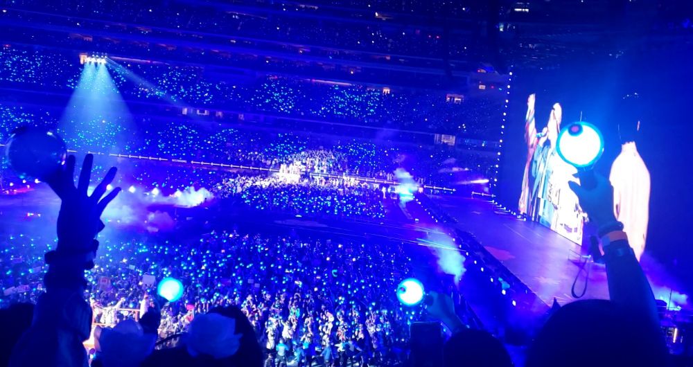 A view of the fourth BTS Permission to Dance on Stage LA Concert. The members are on the jumbo screen to the right and the audience is illuminated by their lightsticks. The image is framed by hands and lightsticks raised in joy and excitement.