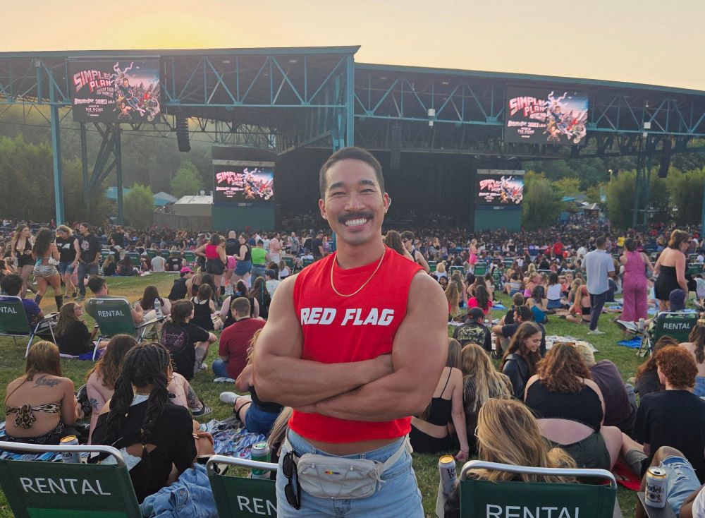 Photo of a man standing in a crowded grassy lawn at the Kesha and Scissor Sisters concert. He is smiling with his arms crossed and wearing a red crop top that says "RED FLAG" in reference to the instant evergreen bop RED FLAG. by Kesha. He is also wearing denim shorts and a fanny pack with a rainbow pin on it bc gay. 