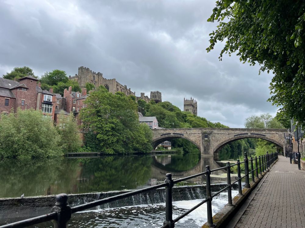 A photo from the riverside path in Durham, showing the River Wear in the foreground, and the castle and cathedral in the distance.  A stone arched bridge is crossing the river, and there are trees on the river bank. The river is calm and reflects the bridge and trees. The sky is cloudy and grey.  