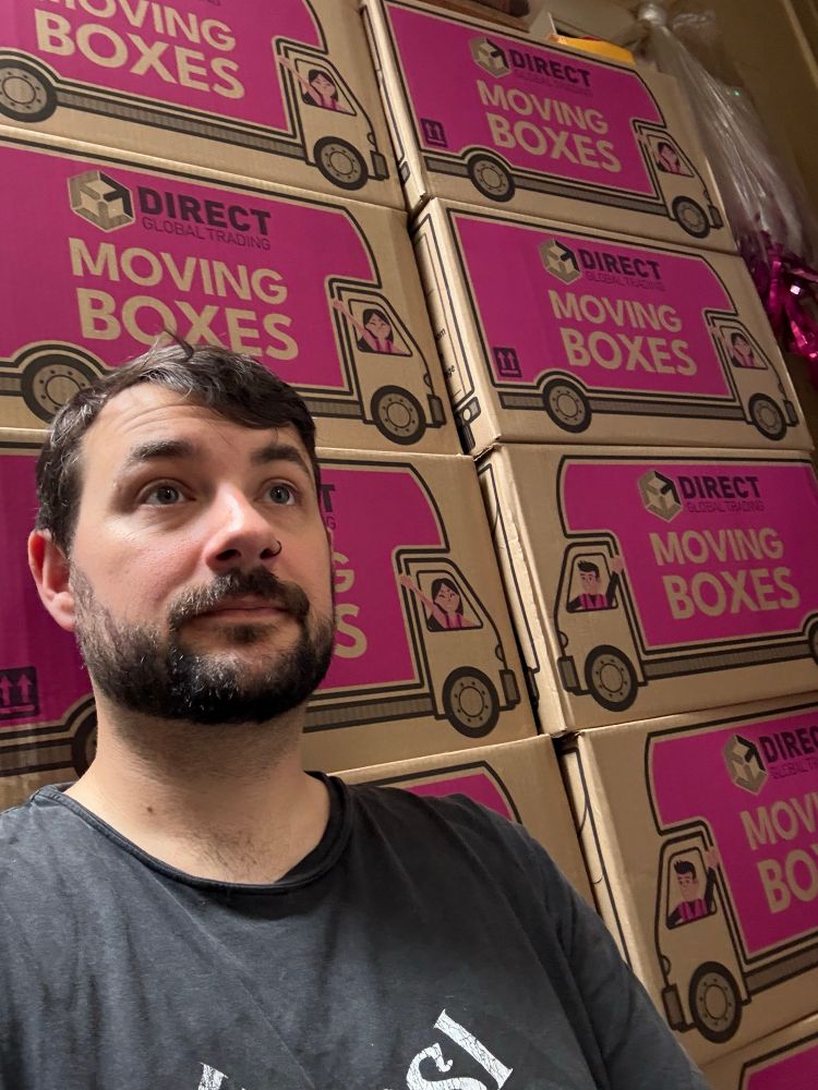 Joe is sitting against a wall of moving boxes with a vacant stare