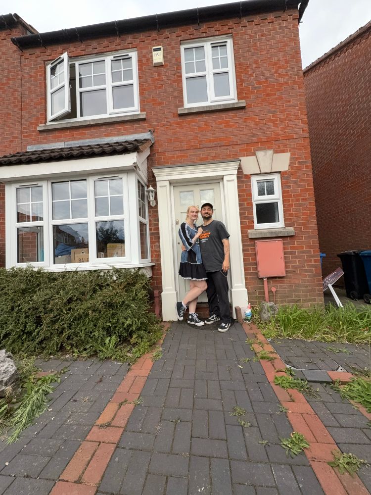 A couple are standing in front of a house with a pale green door and a bush. 