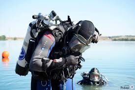 Zwei Taucher*innen. Eine Person steht im Bildvordergrubd, eine Person ist bis auf den Kopf im Wasser