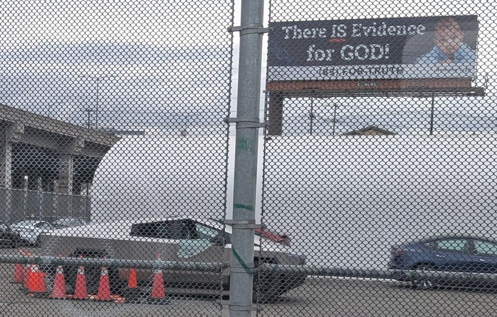 A Cybertruck with a shattered, peeled-down window sits parked surrounded by orange cones. Above it, a billboard reads "There IS evidence for GOD!"
