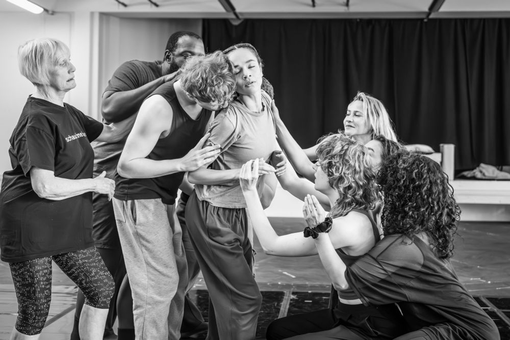 In black and white, a group of people are in movement. In the centre, a man and woman are in an almost embrace while others look on and move towards the action.  

