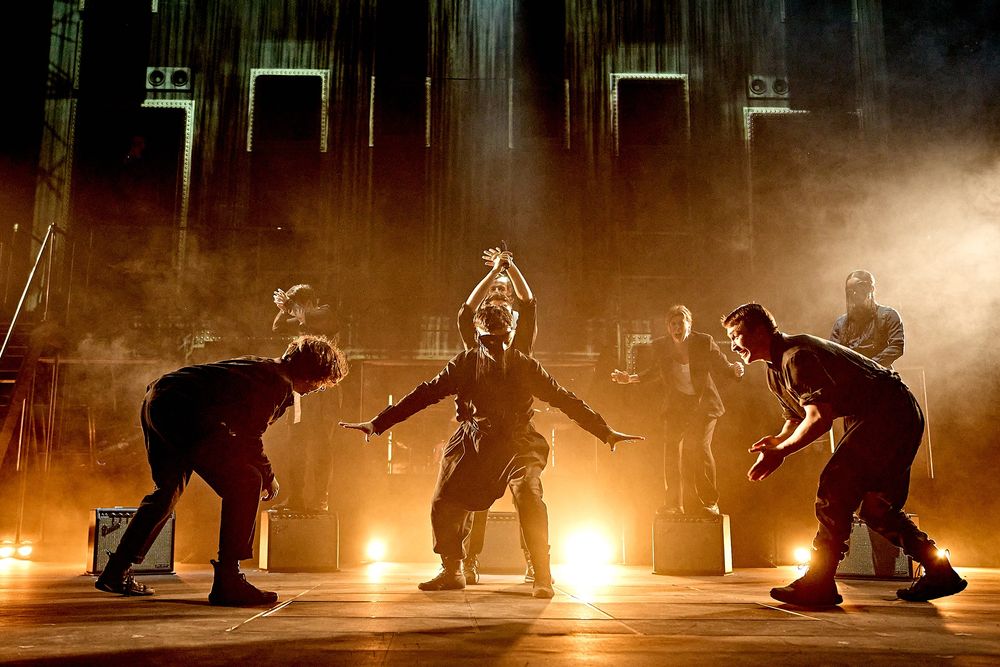 The company of Hamlet Hail to the Thief move dynamically onstage, lit in silhouette by warm-coloured lighting
