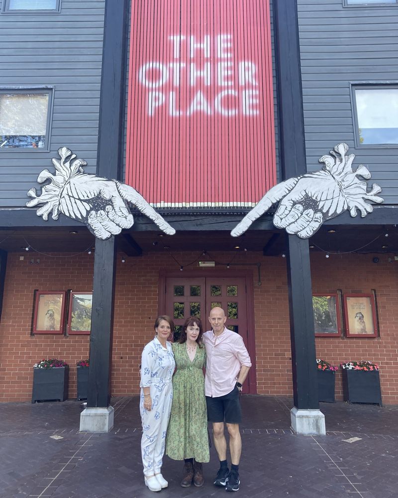 The company of 4.48 Psychosis stand together outside of The Other Place theatre smiling. A tall red sign on the building behind them reads: The Other Place.