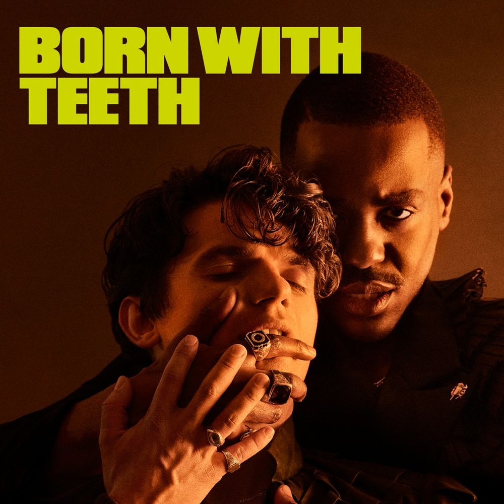 Ncuti Gatwa stands behind Edward Bluemel, with his hand around his mouth. Ncuti looks straight into the camera while Edward has his eyes closed. Behind, a brown faded background. In the top left, Born With Teeth is written in bright green text.