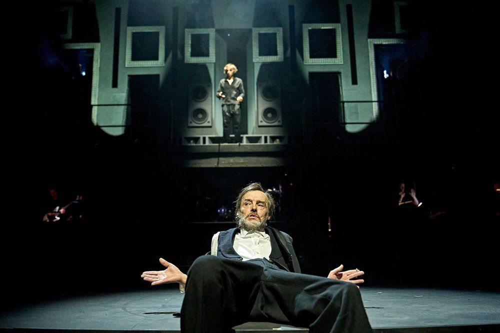 Paul Hilton as Claudius sits and leans back on the front of the stage, wearing black tie. Behind, a figure can be seen lit in the background standing in front of large speaker cabinets 

