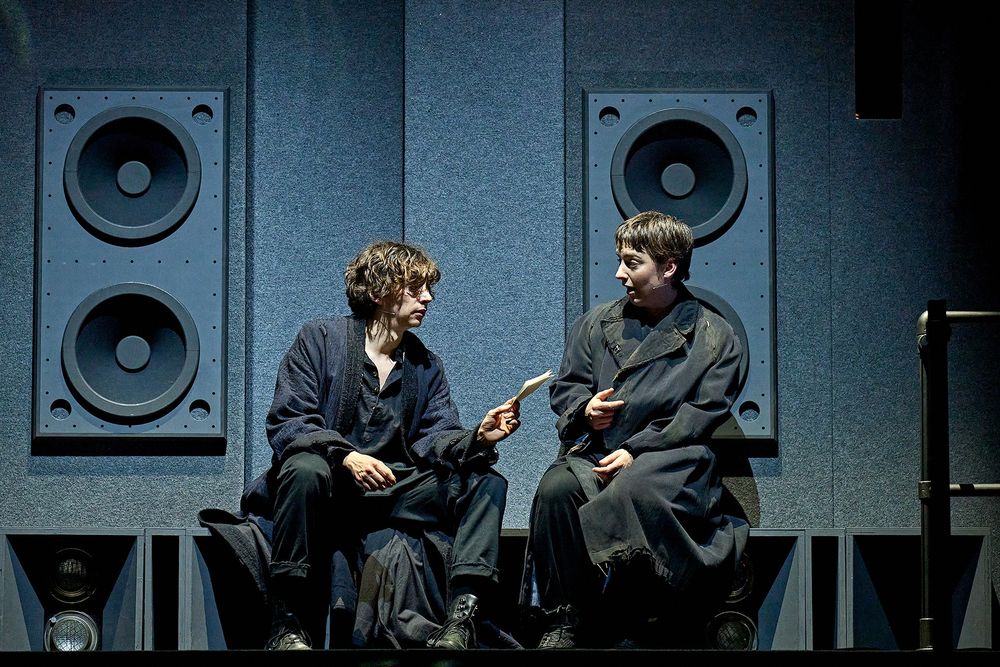 Samuel Blenkin as Hamlet and Alby Baldwin as Horatio sit on a ledge wearing all black. Large speaker cabinets stand behind them. 