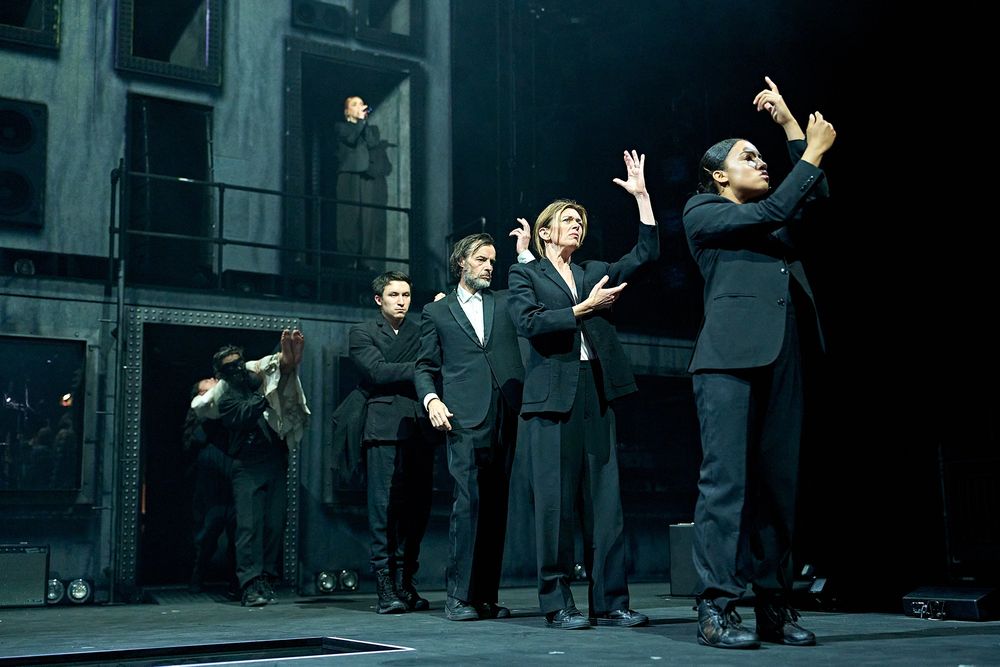 The company of Hamlet Hail to the Thief line up onstage wearing black suits, each gesturing upwards. Behind, speaker cabinets and voids can be seen to the back of the stage