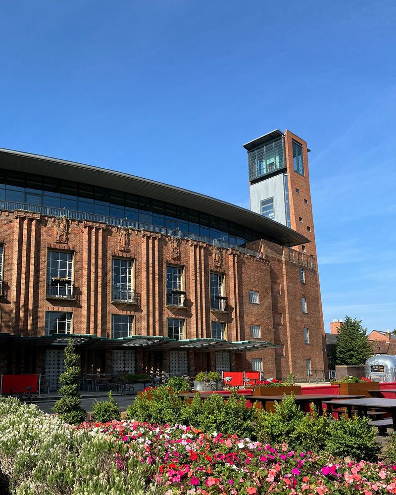 A view of the Royal Shakespeare Theatre from Bancroft Gardens on a summer's day. The red brick building has red, white and pink flowers outside it.