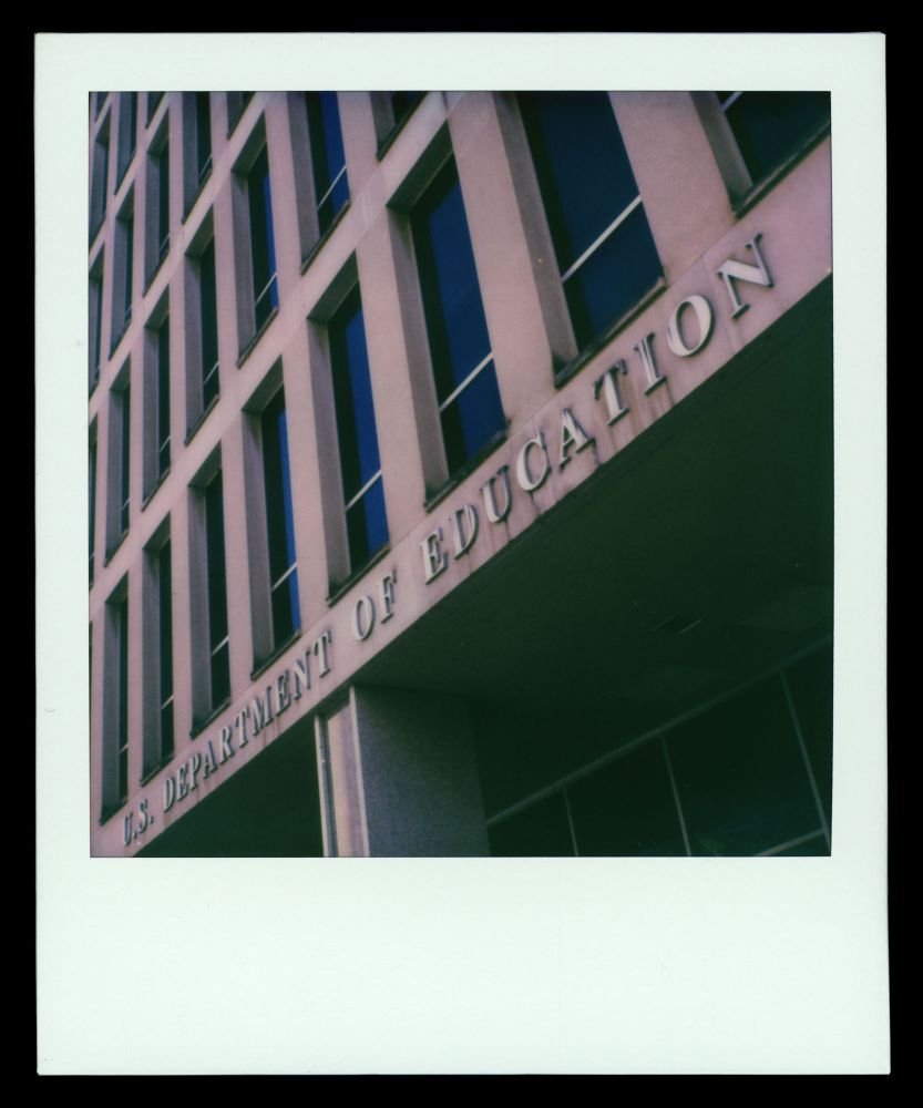 A Polaroid photograph frames an image of a brutralist office building with a masonry grid of tall, rectangular glass windows stretching toward the upper border of the image. The building bears the words "U.S. Department of Education" in slightly-corroded metallic letters affixed to lower portion of the facade. 