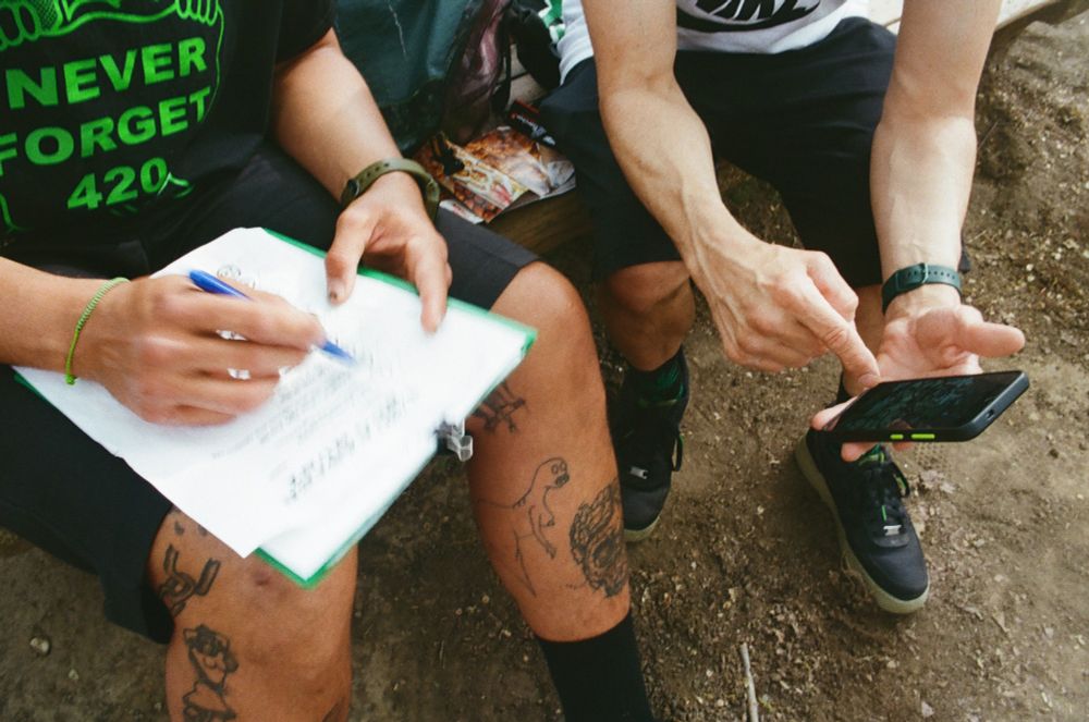 The arms and legs of two people are seen sitting on a bench. One of the people has tattoos on his legs. The other person points at his phone. 