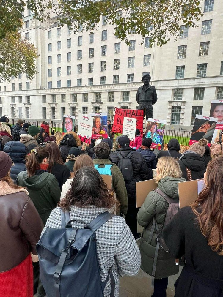 A crowd gathered at a public vigil, holding signs with photos of women, standing near a statue in front of a large government building.
