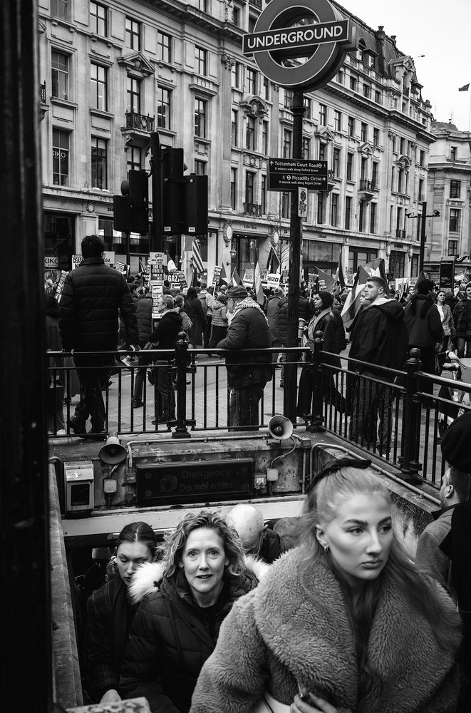 People coming out of the underground at Oxford Circus in London while a march passes in the background.