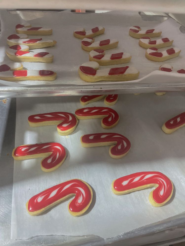 A tray of festive candy cane-shaped sugar cookies, decorated with red and white icing. The cookies on the top rack have a simpler striped design, while the ones on the bottom rack have a more polished marbled look. They are arranged neatly on parchment paper, ready for display or serving.