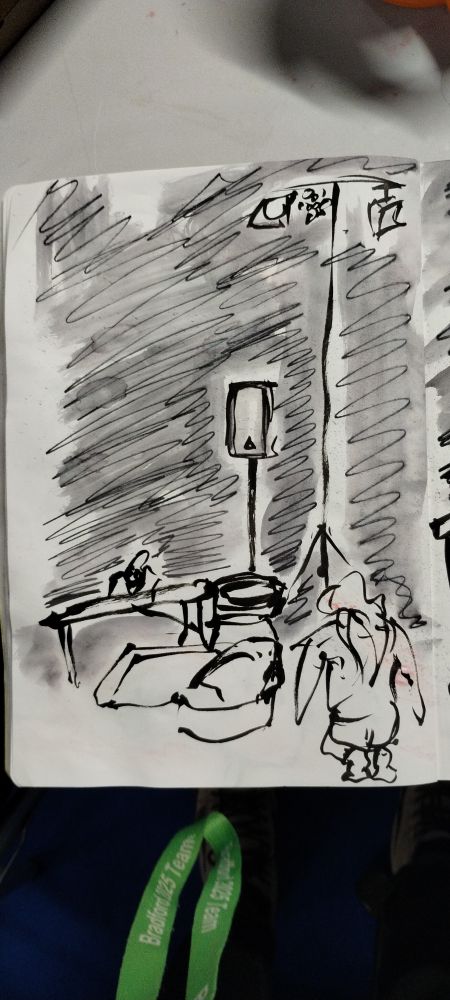 A black and white sketch of actor Beth Organ playing Teagan knelt by a suitcase. In the background is a stand with a lights and a stand with a speaker and Tom doing lighting behind his desk