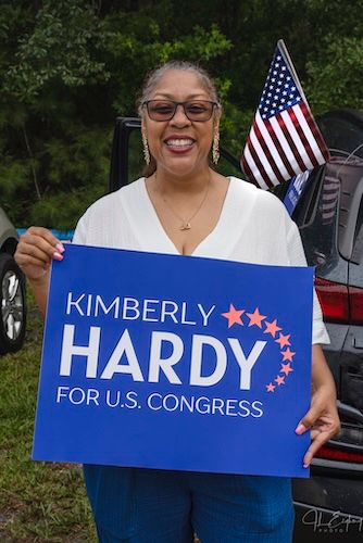 Kimberly Hardy, in a white top and jeans, stands outside at a community event on a summer day, holding a sign that says "Kimberly Hardy for US Congress." There's a US flag in the background. 
