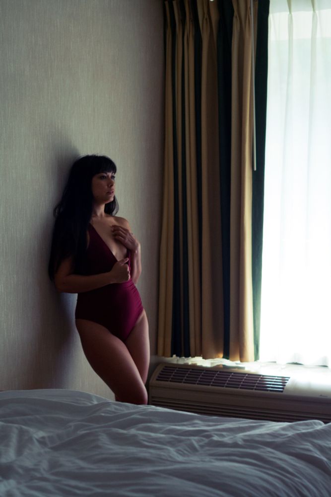 Profile of a woman leaning up against a wall, between a bed and air conditioning unit in a hotel room. She is wearing a burgundy bodysuit, pulling it open and covering her breast with her left hand.
