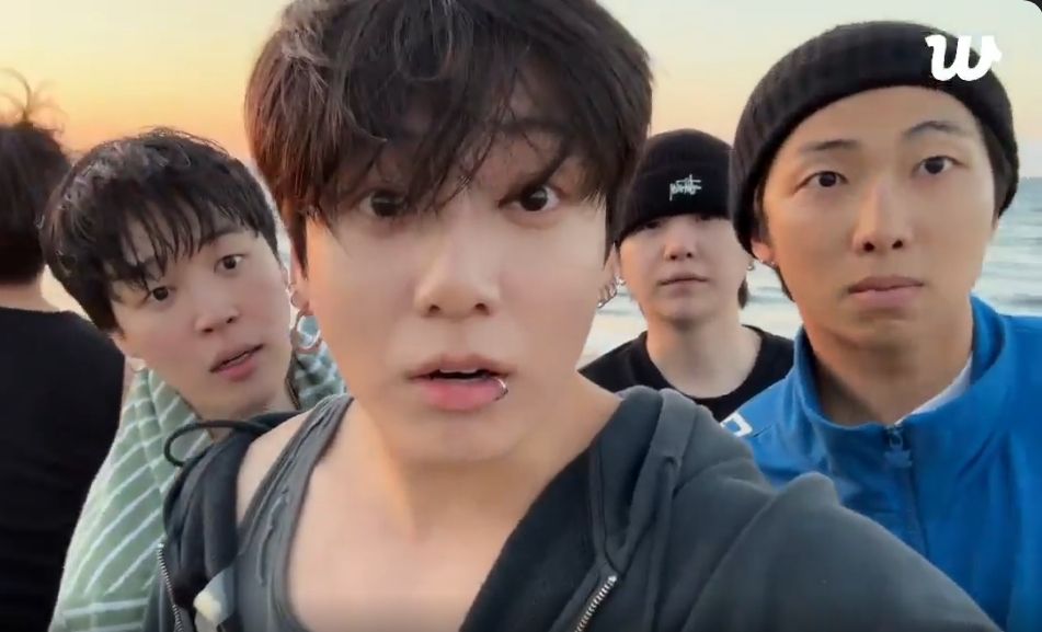 BTS at the beach in LA. Jungkook is center with a surprised and puzzled look on his face. Around him are Jimin, Yoongi, and Joon, all looking at the camera with seriously expressions. The back of Tae's head is visible. Hobi and Jin are also there, but not pictured.