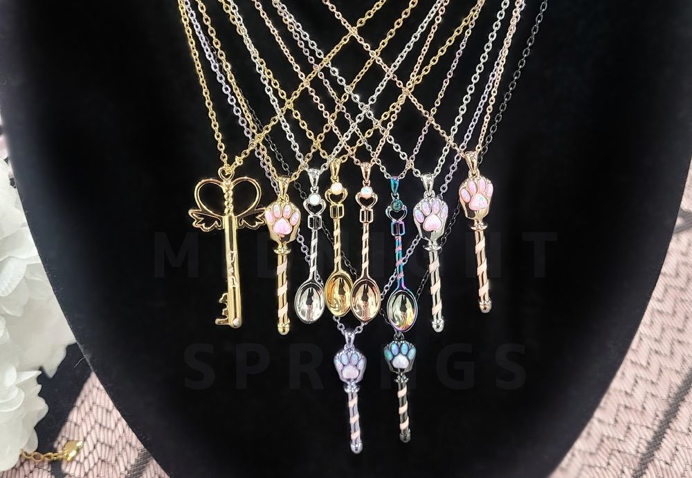 Velvet jewelry display with almost all of my necklace designs pictured; a golden magical girl-inspired key, cat paw-inspired wands in rose gold, gold, silver, purple, and black with glittery enamel spiraling up the rod portion of the design, and spoons in silver, rose gold, gold, and rainbow to represent the "spoon theory" with the same spiraling enamel feature.