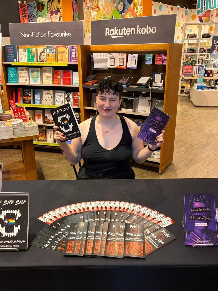 A photo of Pride with a Bite Co-Founder Sarah Laudenbach behind her signing table with copies of "We Bite Back" and "Through the Realm of the Vacant Prince."
