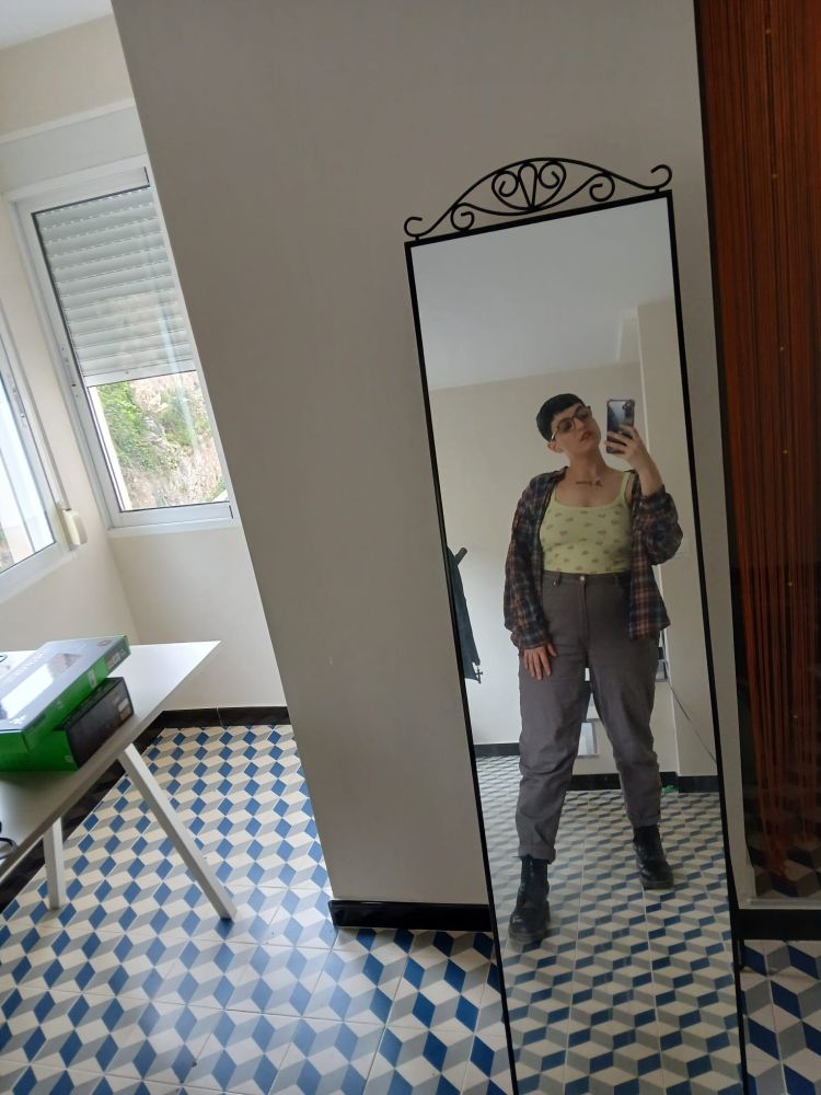 Foto mía en el espejo de casa de mi novia y su novia. Llevo unos pantalones vaqueros largos de talle alto, ceñidos a la cintura, color marrón oscuro; una camiseta corta verde claro con estampado de fresitas color rosa, de tirantes, ceñida; una camisa de cuadros oscuros abierta por encima y botas militares negras. Llevo también gafas de pasta oscuras y ovaladas y algo de pintalabios rojizo.

Tengo el pelo castaño oscuro, liso, muy corto, con flequillo recto corto. Llevo un tatuaje bajo las clavículas, sobre el pecho, que dice A jierro en tinta negra, en letra gótica. Apoyo una mano sobre el muslo y con la otra sujeto el móvil haciendo la foto. Estoy seria, tengo la boca entreabierta y miro hacia abajo de lado.

El espejo es de pie, alto, rectangular, con marco de forja negra ornamentada en la parte de arriba. Está apoyado en la pared, blanca. Al lado se ve una cortinilla de hilo naranja. El suelo tiene patrones geométricos azules, blancos y grises. Se ve también parte de un escritorio blanco con cosas apoyadas encima y unas ventanas cerradas, con la persiana a medio bajar, por las que entra mucha luz natural.