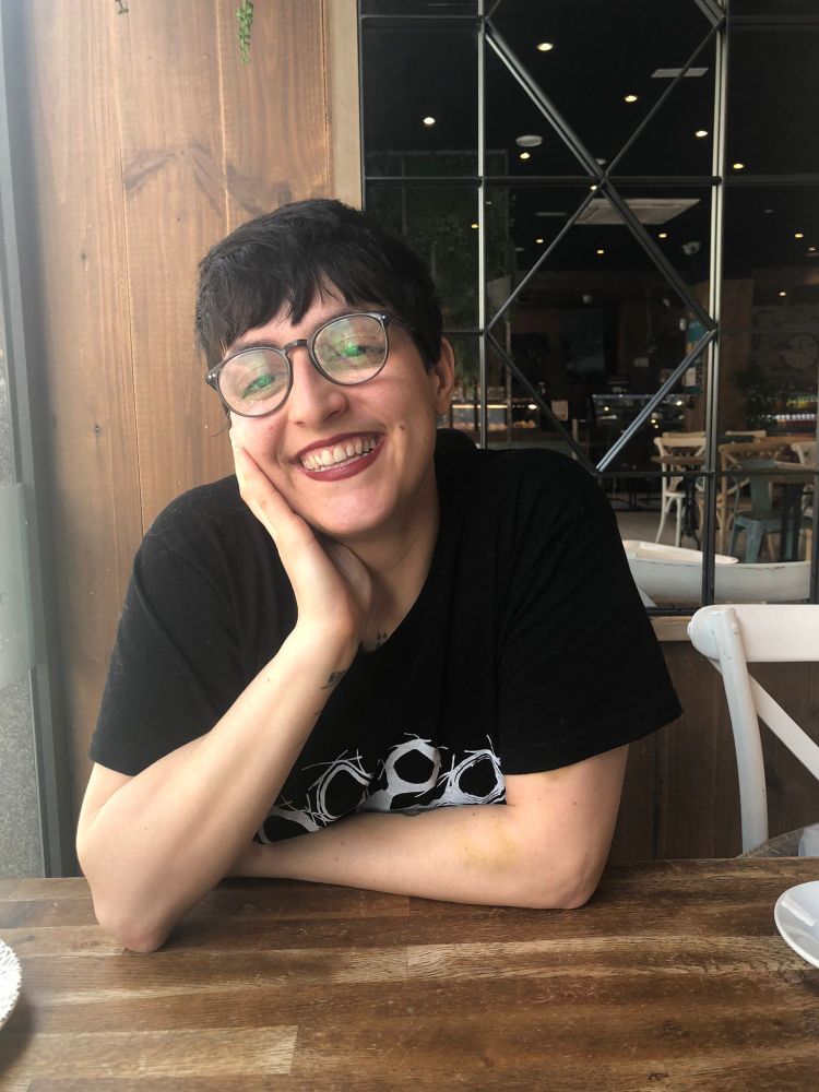 Foto mía, lesbiana cansada sentada en una cafetería mirando a la cámara con mucha luz diurna entrando por la ventana de mi derecha. Tengo el brazo derecho en la mesa de madera oscura y el brazo izquierdo doblado por el codo, sujetando la cara con la mano. Se entreven cachitos de tatuajes en la muñeca derecha y bajo el cuello. Tengo los ojos un poco entrecerrados y sonrío con la boca abierta.

Tengo el pelo castaño oscuro liso, muy corto, con flequillo despeinado; y ojos color marrón oscuro. Estoy un poco pálida.

Llevo una camiseta negra de manga corta algo ancha en la que se entrevé el logo de Tribade en blanco en el centro, gafas oscuras grandes ovaladas y pintalabios granate oscuro mate.

Detrás de mí, un panel de madera oscura de la cafetería, mesas oscuras y sillas blancas al fondo y muchas luces en el techo.