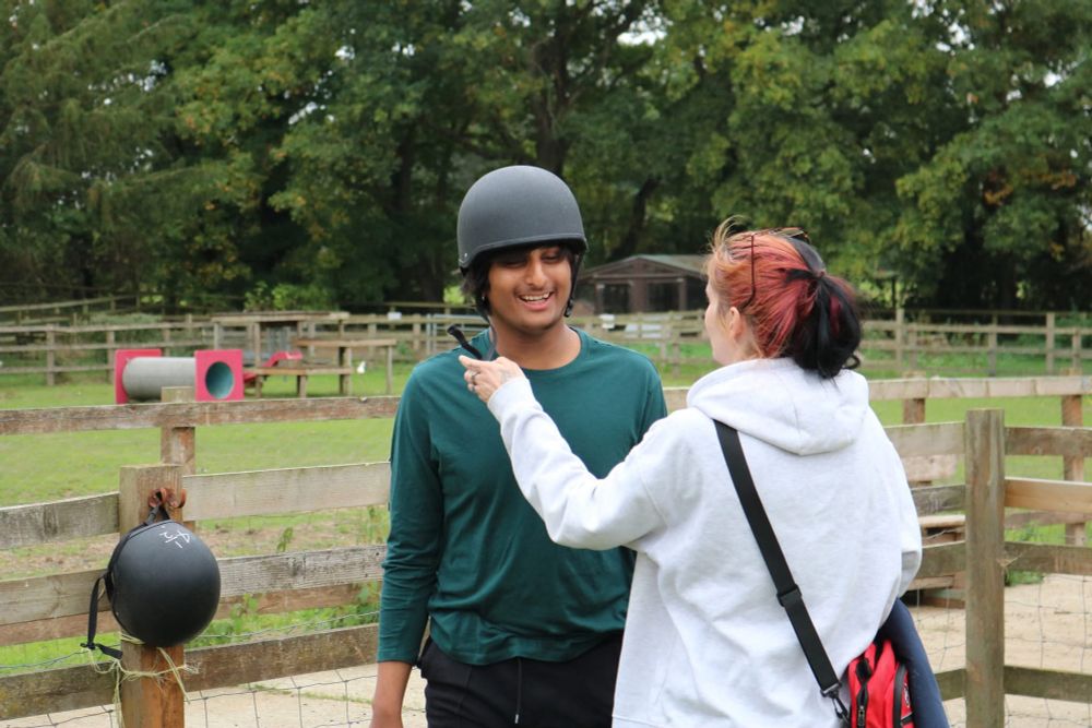 Photo of a boy, laughing. He is wearing a green top and has a black riding helmet being put on him by a lady in a white hoodie. 