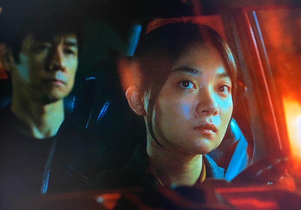 A young Japanese woman looking in the rearview mirror at an older man in the backseat. 