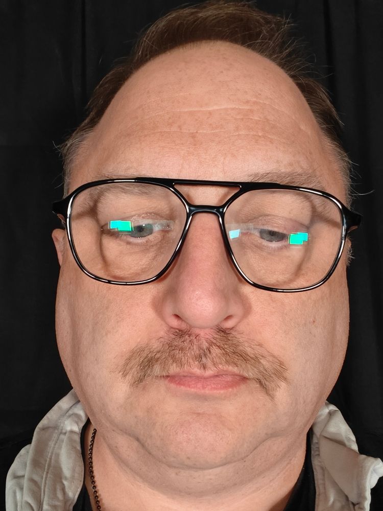 A middle aged white man's moon face with glasses. On his top lip a rag-tag group of hairs are coming together against all odds to form a mustache.