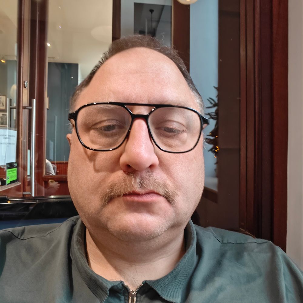 A moon-faced middle-aged white man with glasses showing off his mustache.