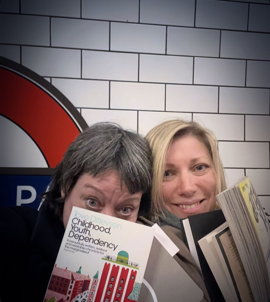 Helen Macdonald and Elvira Lind at Holland Park tube station posing with books in the white glare of platform lights. White tiles with black grout behind, Helen holding a copy of CHILDHOOD, YOUTH, DEPENDENCY 