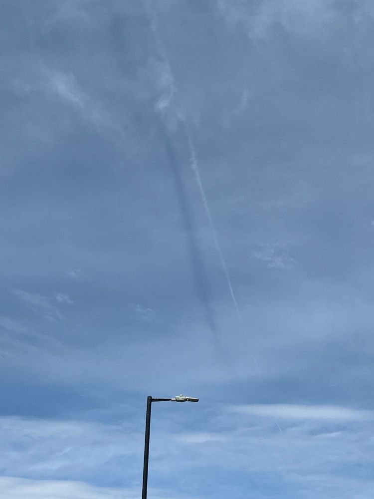 High aircraft contrail with a darker line below it that is the contrail’s shadow on a layer of thin cloud. In the mid ground, a streetlamp