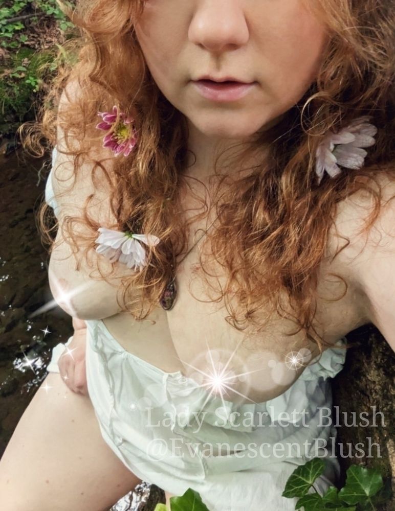 Photo of me with my dress pulled down to show my breasts and flowers in my hair next to a creek in the woods.