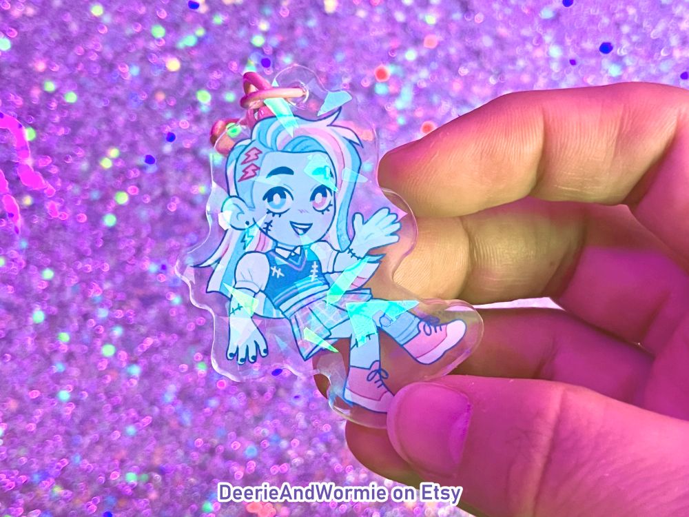 a photo of my hand holding a trans colors frankie stein keychain with a broken glass holo finish with text that says "DeerieAndWormie on Etsy"