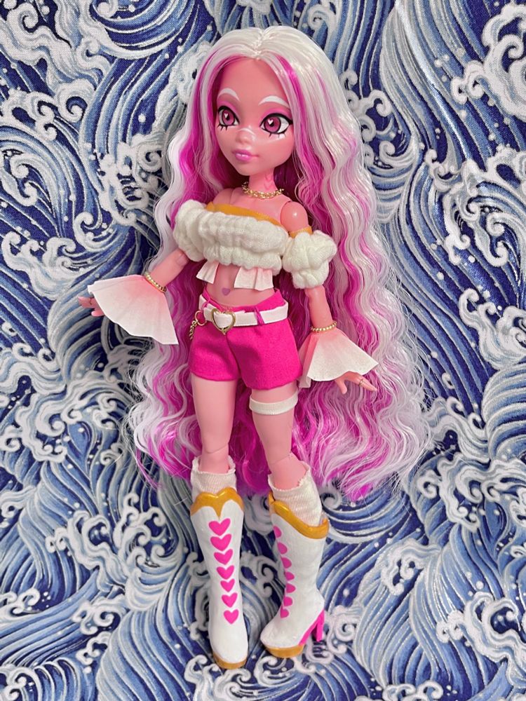 a photo of a customized monster high doll of a pink triton dnd character. she has long wavy white hair with magenta on the underside, frilly fins on her ribs and wrists, and she is wearing a puffy white off-the-shoulder top with magenta shorts and white boots with pink heart designs, and lots of gold jewelry
