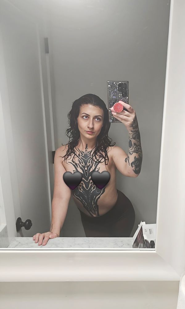An alternative brunette girl taking a mirror selfie with no shirt on and her torso tattoo on full display with her breasts censored. 