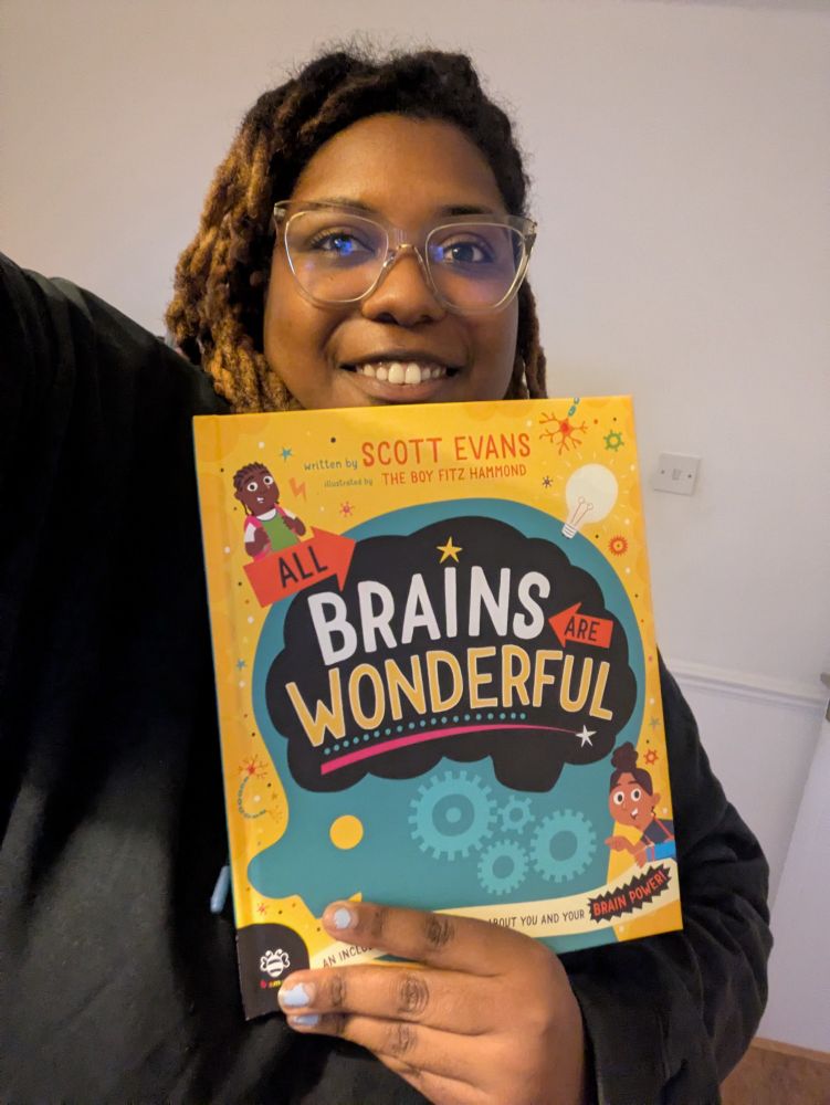Rachel poses for a selfie with a book titled All Brains Are Wonderful. The cover is bright yellow with an illustration of the silhouette of a head in blue, with the title written against the black silhouette of a brain.