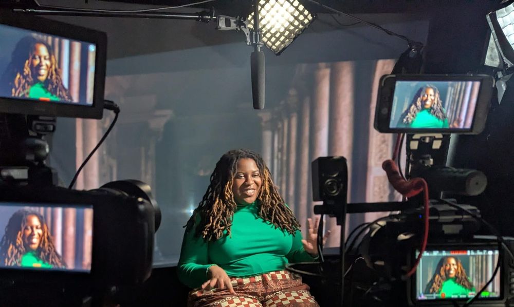Rachel is wearing a green top and sits on a chair on set in a studio. She is surrounded by 4 screens showing different camera angles of her while she speaks.