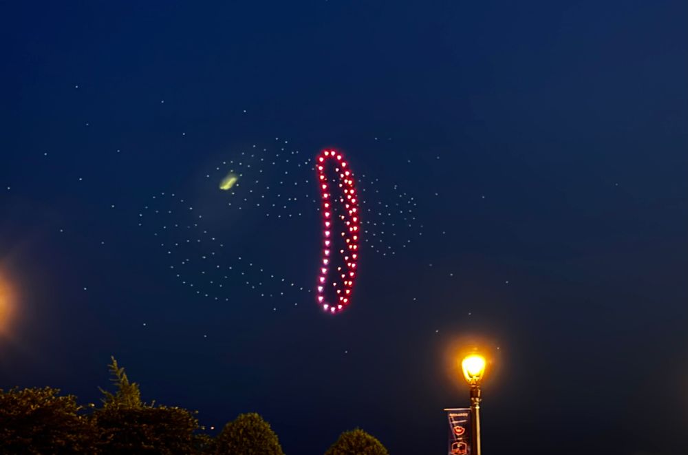 A sausage made of illuminated drones floats in the evening sky. 