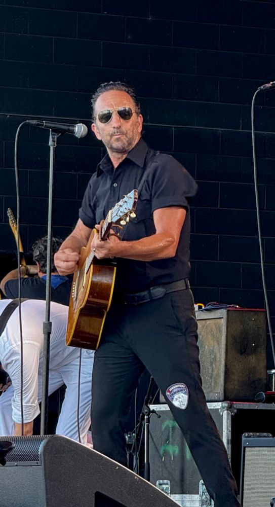 Photo of a man (Ike Reilly) wearing sunglasses and playing acoustic guitar 