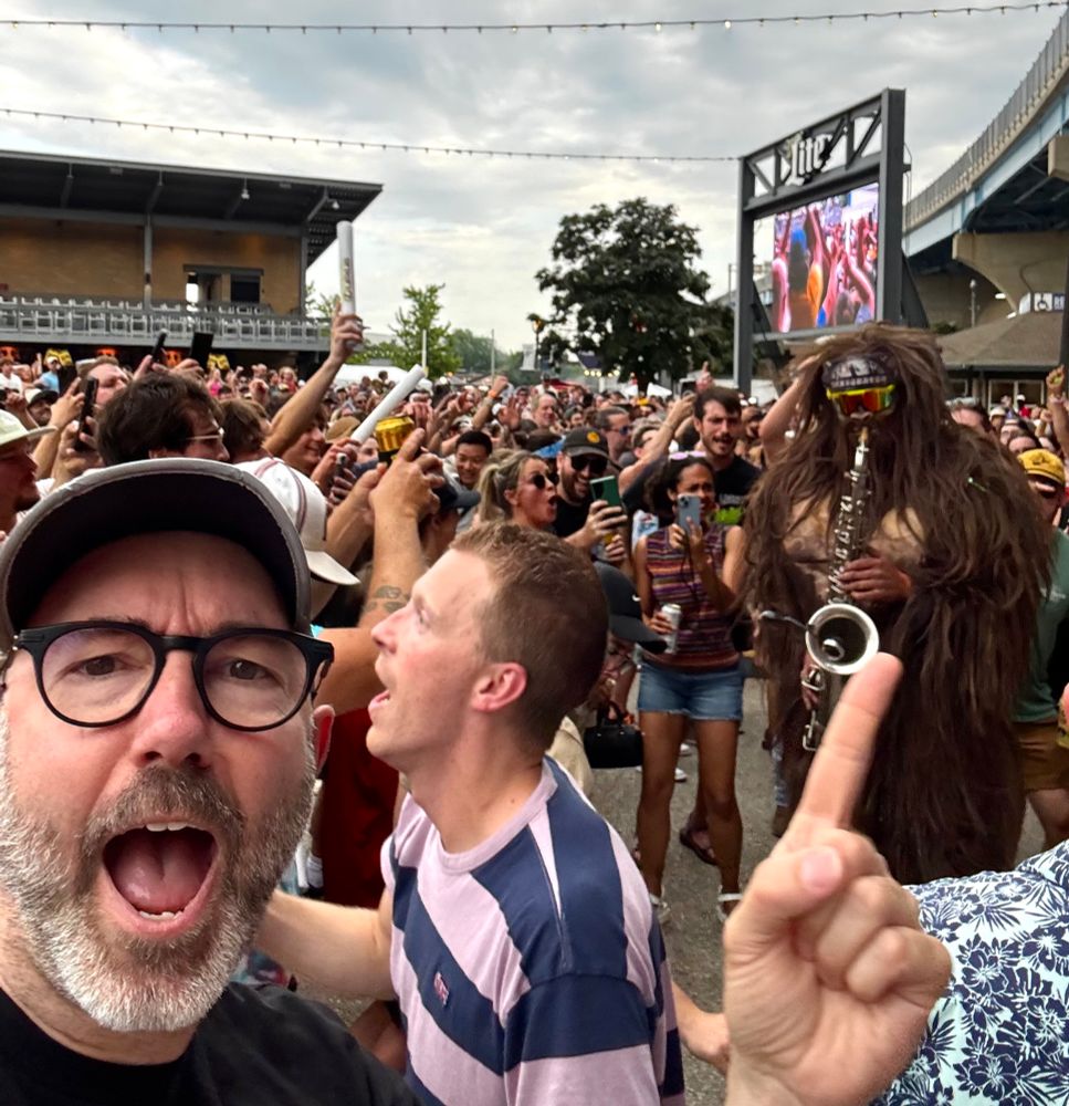 Photo of a Sasquatch playing saxophone in a crowd with a handsome, bearded man in the foreground pointing at the Sasquatch. 