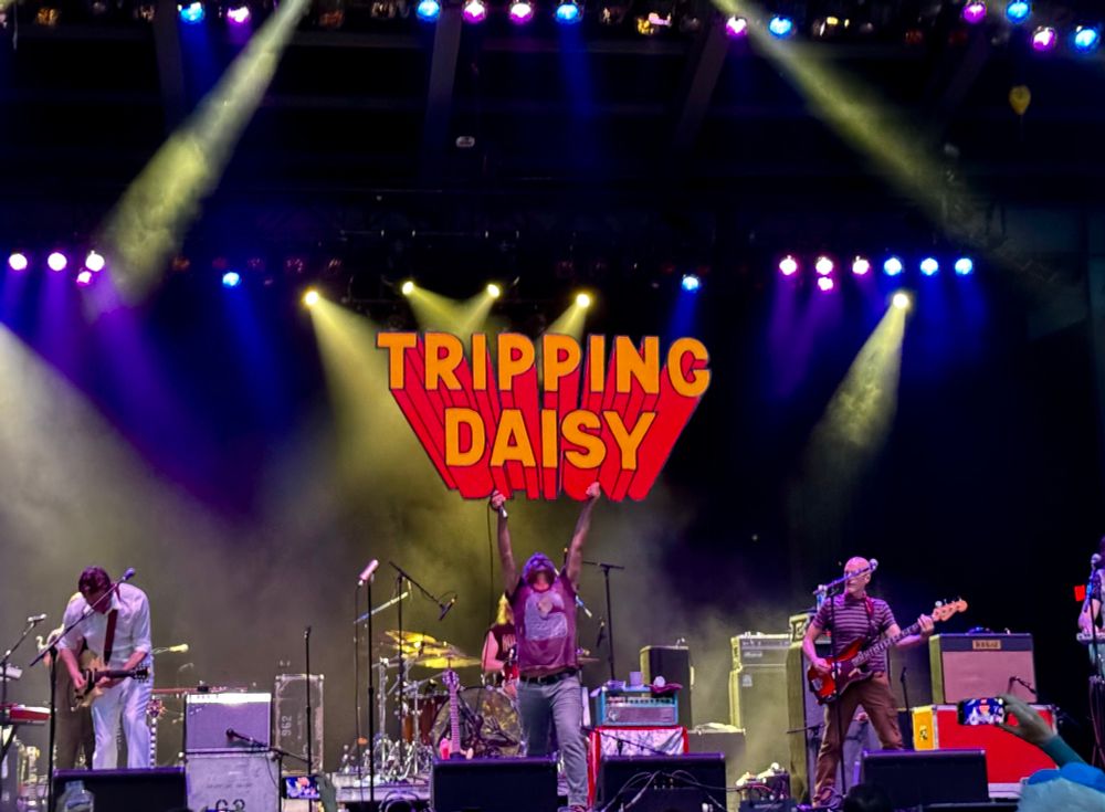 Live performance of the band Tripping Daisy. Singer Tim DeLaughter is holding an orange & red sign that says, “tripping Daisy”above his head. 