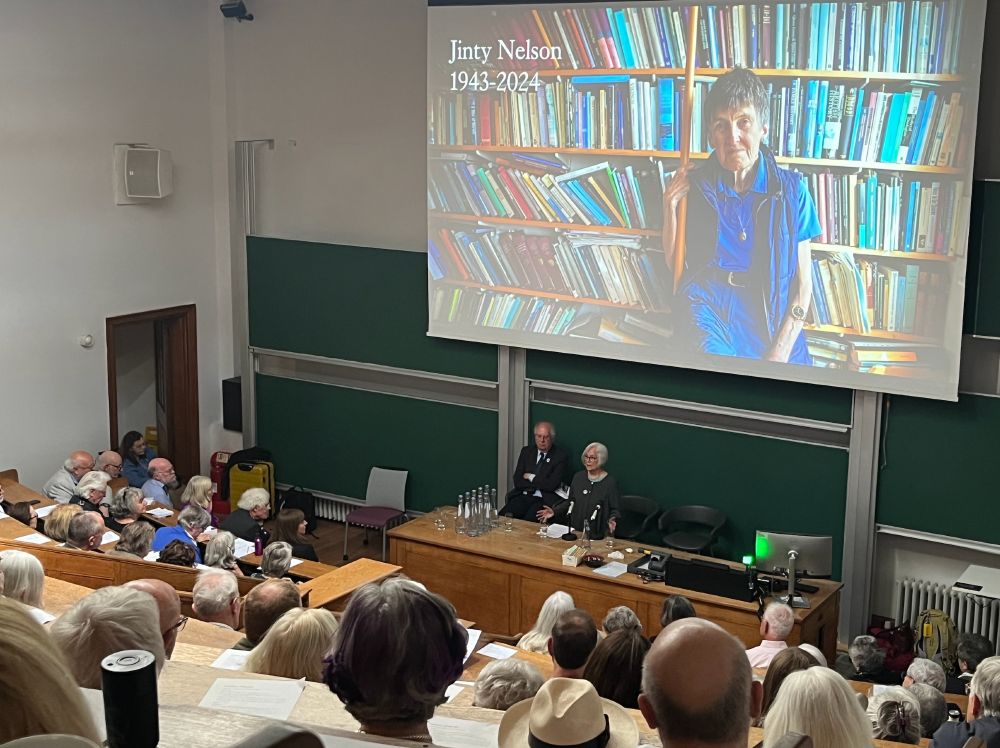 Attendees of the Jinty Nelson memorial event at King's College London with a large banner image of Jinty Nelson. Speakers are Pauline Stafford and Peter Mandler.