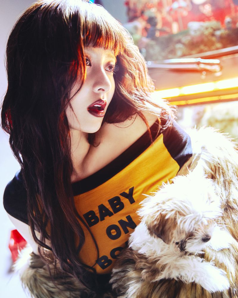 Huh Yunjin of Le Sserafim in a black wig with baby bangs and dark makeup and wearing a yellow shirt that says “Baby on Board” and a white furry jacket for the “Cheeky Neon Pepper” concept photos for Le Sserafim’s Upcoming comeback “Spaghetti” 